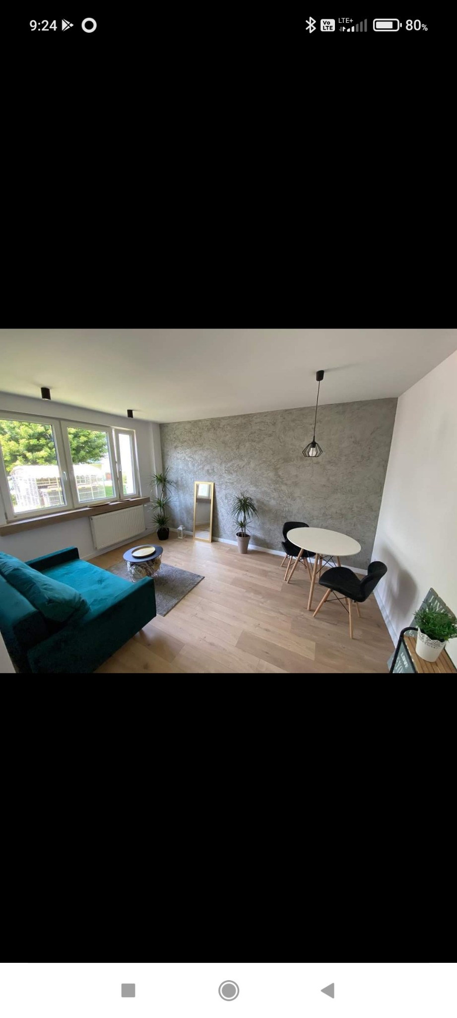 Jasny salon z drewnianą podłogą, turkusową sofą, okrągłym stolikiem kawowym, białym stołem z czarnymi krzesłami, dużym oknem i ścianą w odcieniach szarości z dekoracyjnym tynkiem.