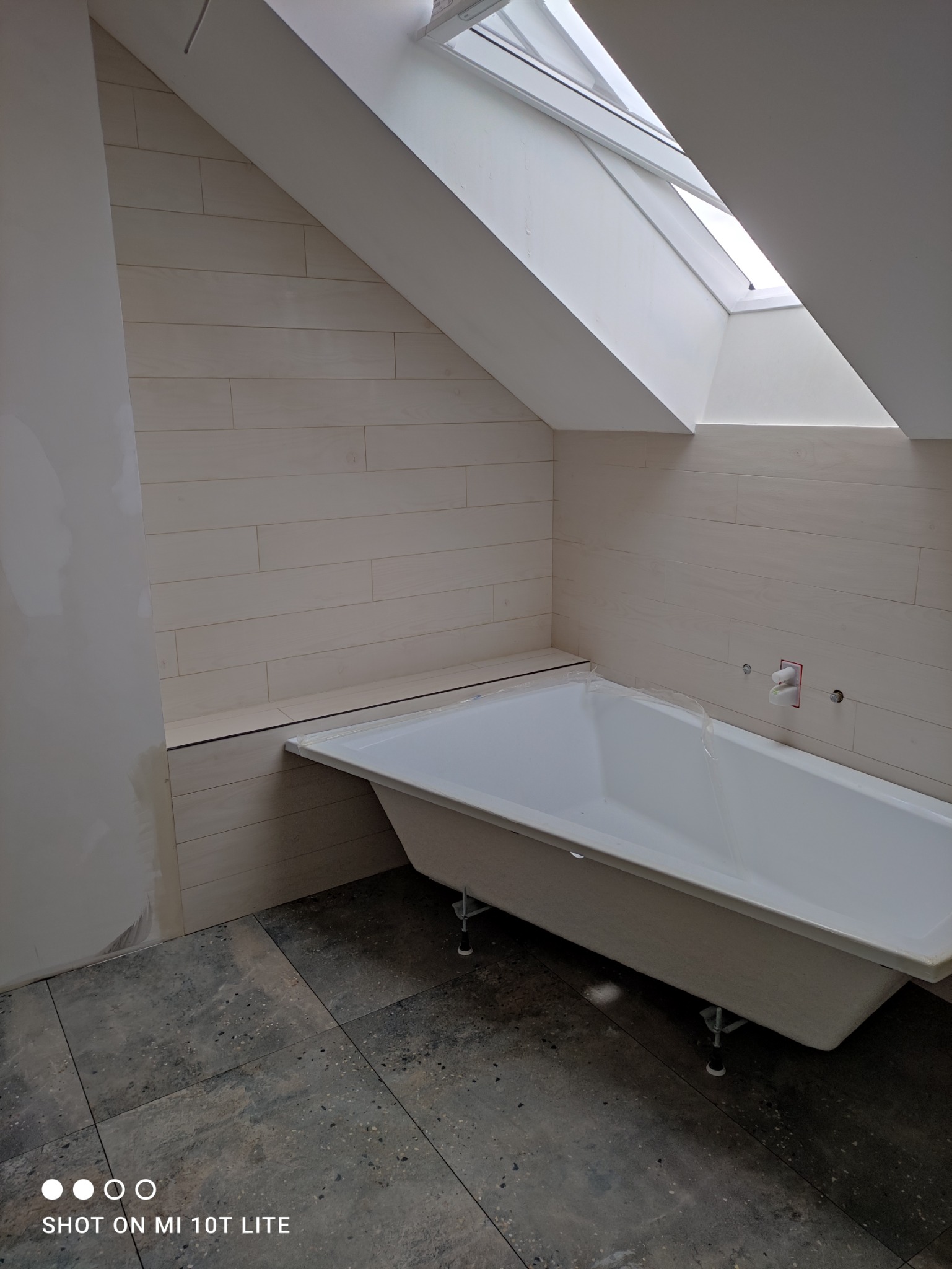 Wanna w łazience na poddaszu, ściany wyłożone płytkami imitującymi drewno, widoczne okno dachowe i instalacja wodna przed podłączeniem baterii.