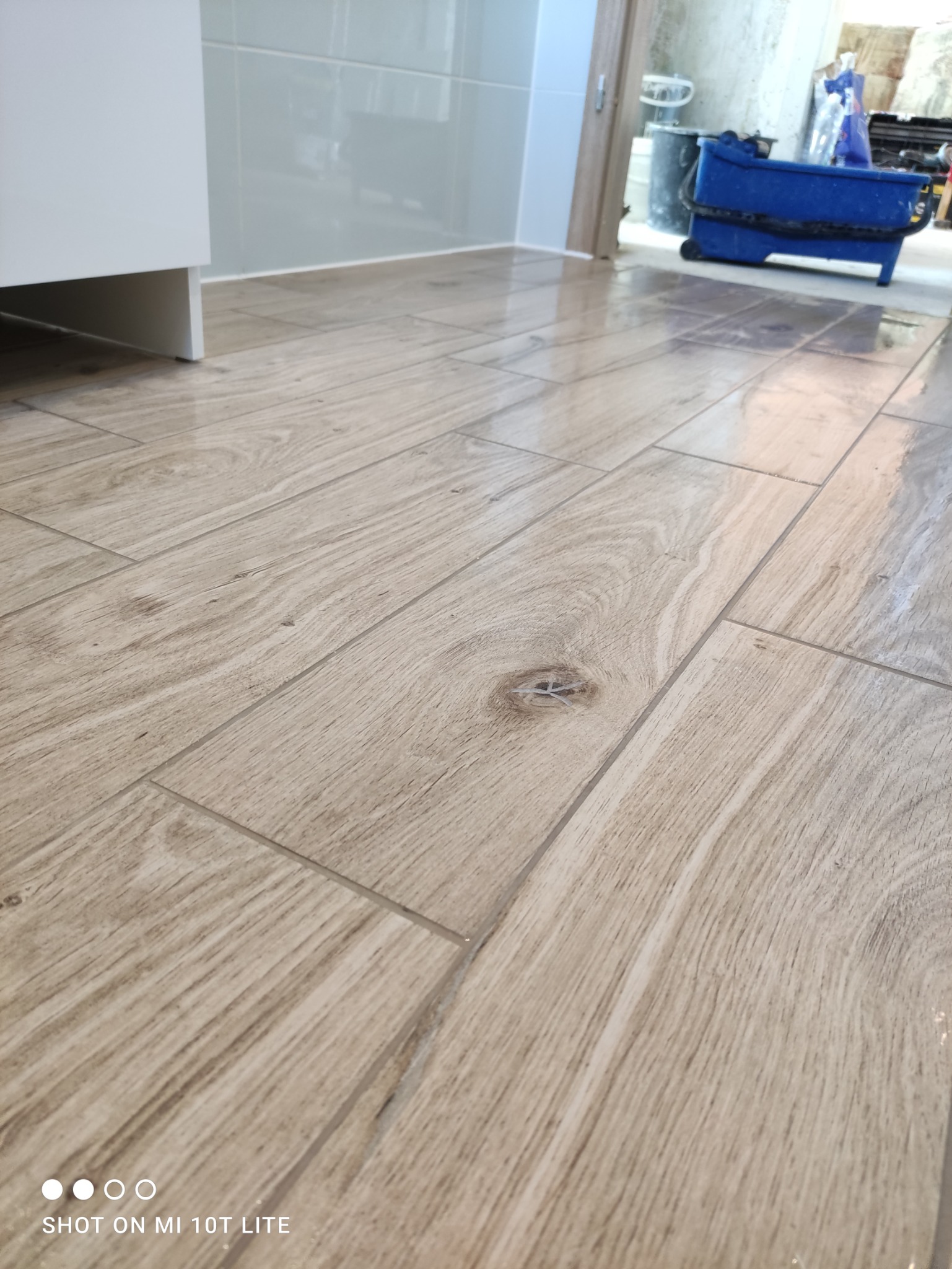 Ułożona podłoga z płytek imitujących drewno w łazience, widoczny fragment szafki pod umywalkę i wiadro z akcesoriami w tle.