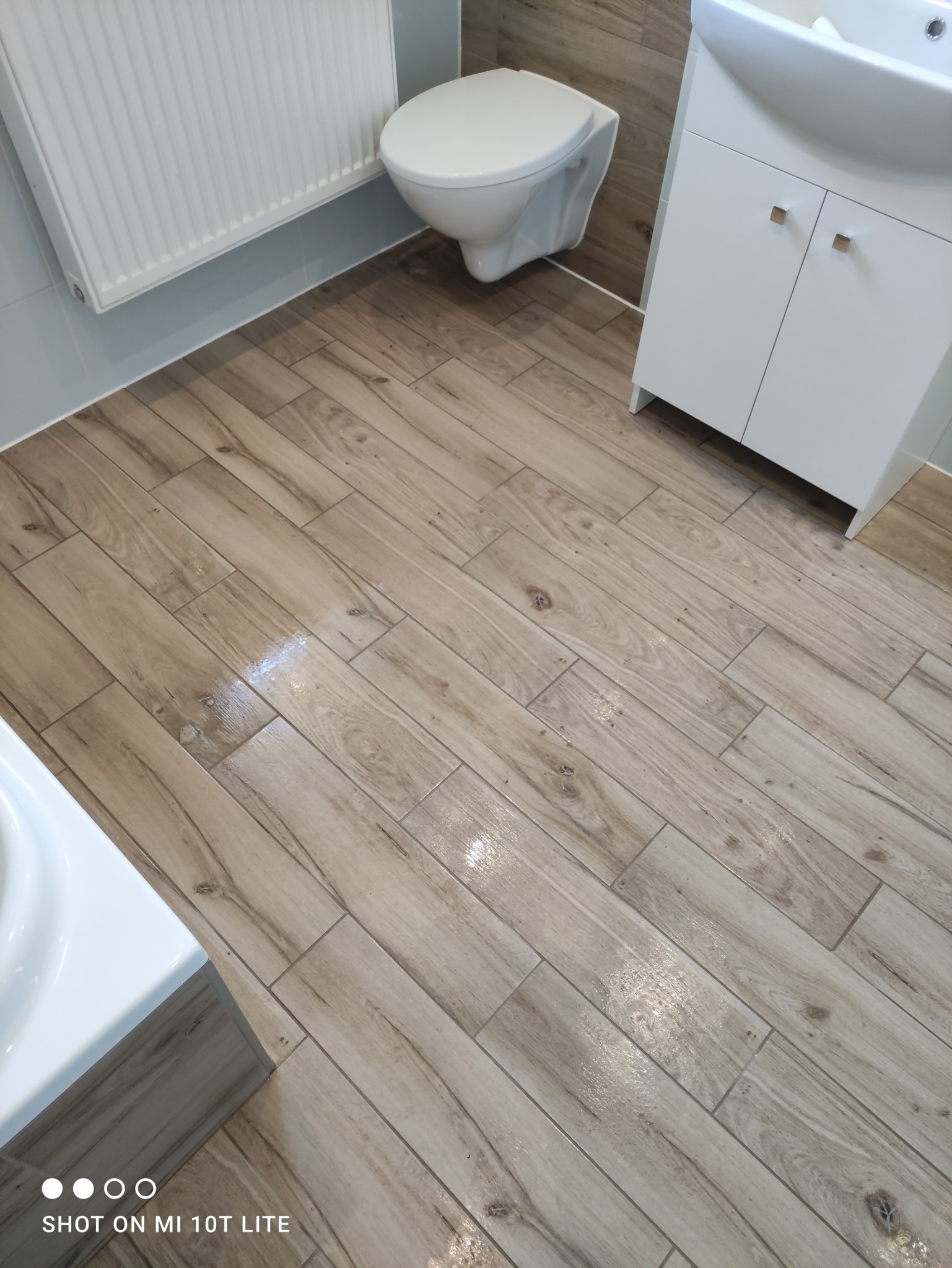 Jasne, drewnopodobne płytki podłogowe ułożone w łazience z podwieszaną toaletą, grzejnikiem drabinkowym i białą szafką pod umywalką.