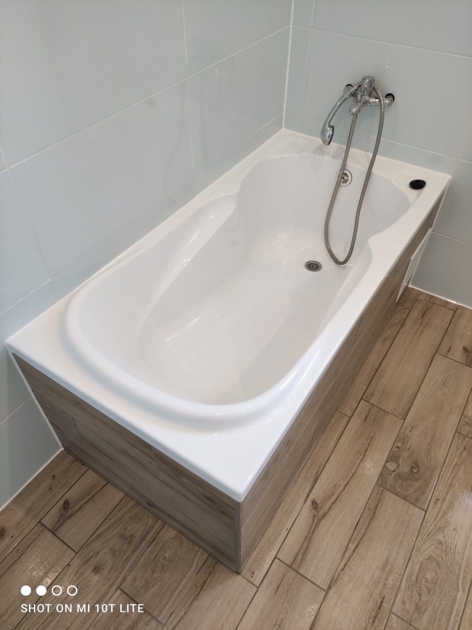 Biała wanna obudowana panelami imitującymi drewno, chromowana bateria z prysznicem, jasne płytki na ścianach i podłodze z wzorem drewna.