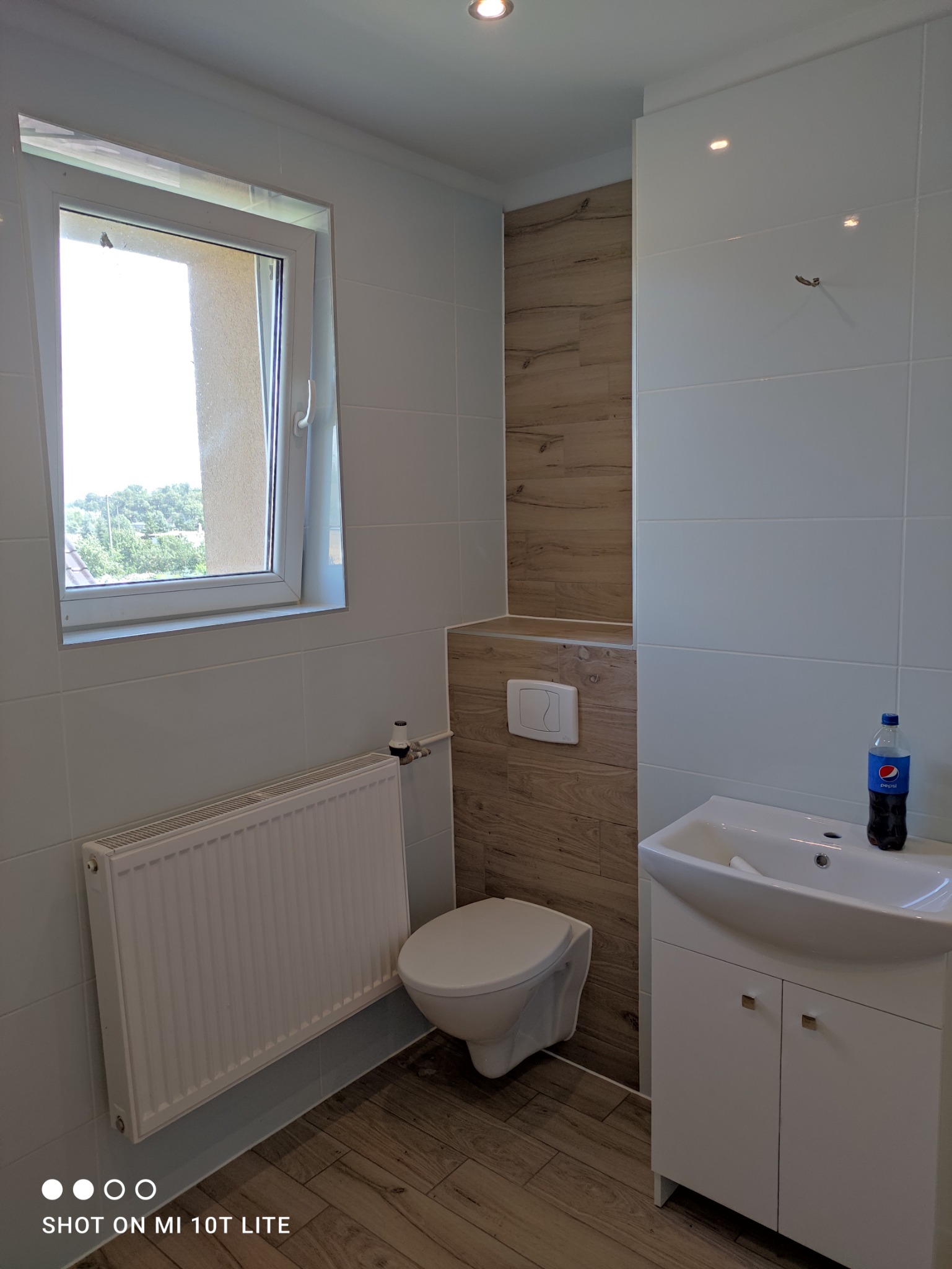 Wykończona łazienka z białymi płytkami, drewnopodobnymi akcentami, podwieszaną toaletą i białą szafką z umywalką, na której stoi butelka Pepsi.
