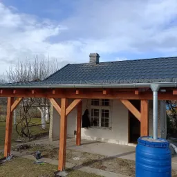 Nowa, ciemna blachodachówka z rynnami na drewnianej konstrukcji z czterema słupami, przybudowanej do ściany budynku z oknem i niebieskim zbiornikiem na deszczówkę.