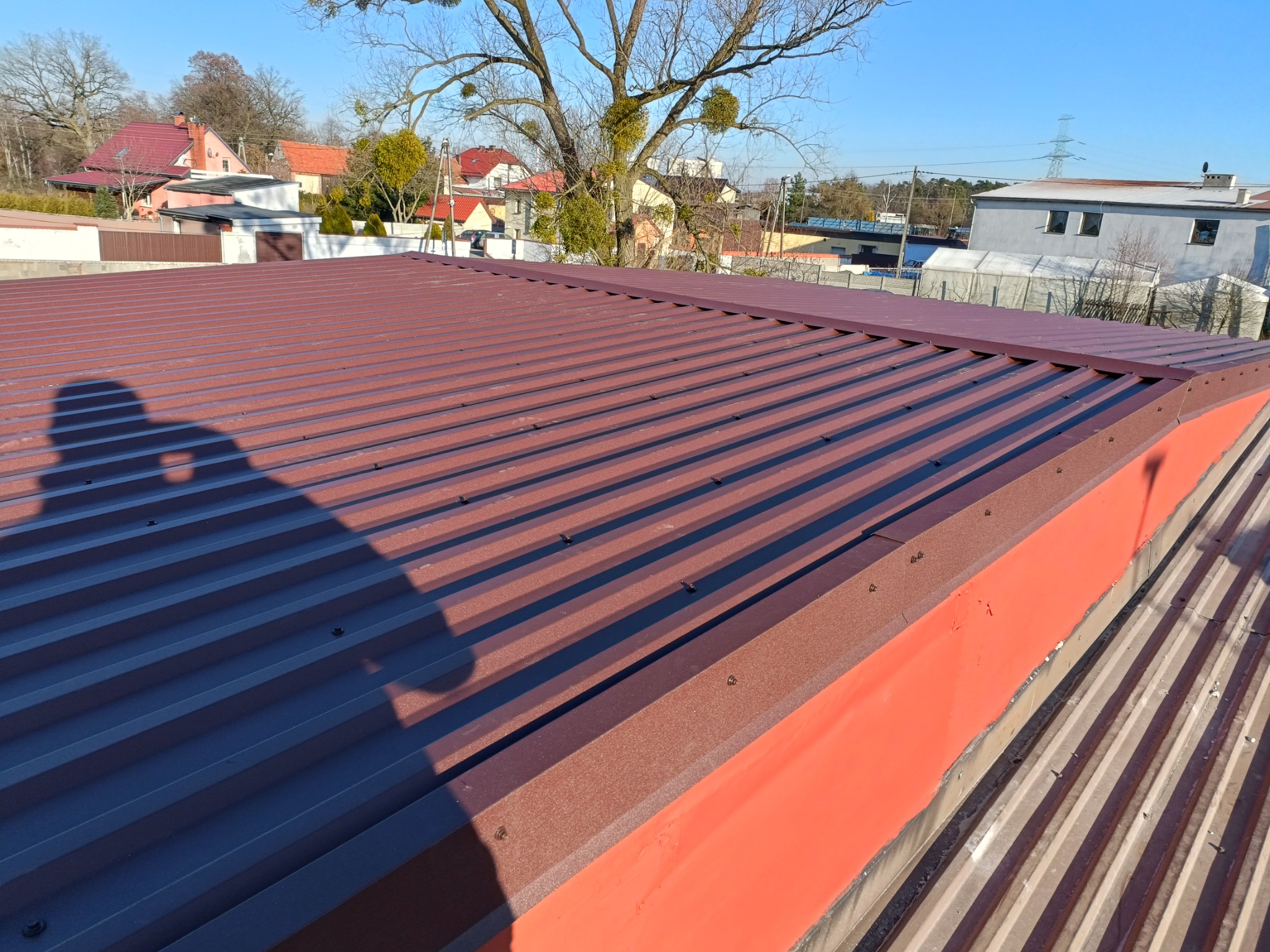 Nowy, brązowy dach z blachy trapezowej, widoczny z góry, z cieniem osoby fotografującej, w tle budynki mieszkalne i drzewa bez liści.