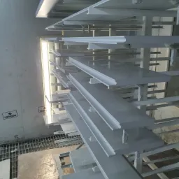 Elementy wykończeniowe, prawdopodobnie listwy przypodłogowe, świeżo pomalowane na szaro, wiszące na metalowym stojaku w lakierni z widocznym systemem wentylacji w tle.