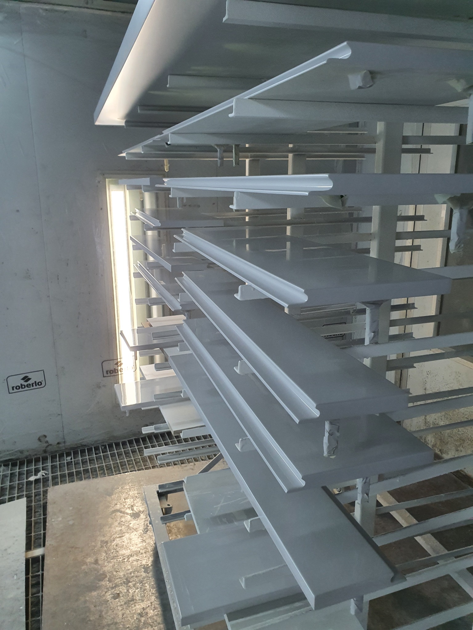 Elementy wykończeniowe, prawdopodobnie listwy przypodłogowe, świeżo pomalowane na szaro, wiszące na metalowym stojaku w lakierni z widocznym systemem wentylacji w tle.