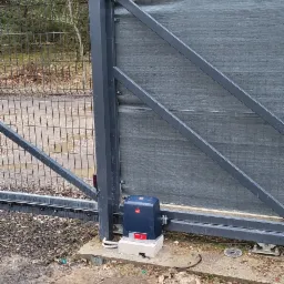 Zamknięta brama przesuwna z napędem elektrycznym i lampą ostrzegawczą, widoczna na tle drzew.