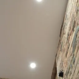 Dwa okrągłe, punktowe źródła światła LED w suficie podwieszanym z drewnianą listwą, ściana z cegły w tle.