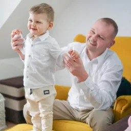 Uśmiechnięty ojciec pomaga synkowi w białej koszuli i beżowych spodniach wstać z żółtej pufy. Chłopiec patrzy w lewo z radością.