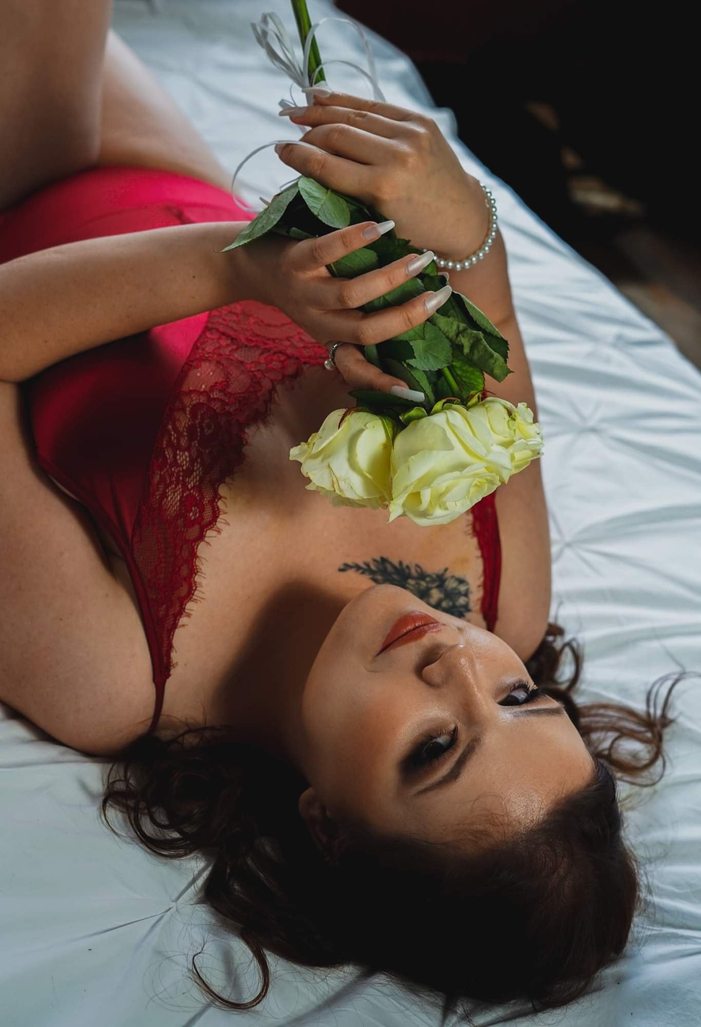 Kobieta w czerwonej bieliźnie leży na białej pościeli, trzymając żółte róże w dłoniach z długimi paznokciami, widoczny tatuaż na dekolcie i perłowa bransoletka na nadgarstku.