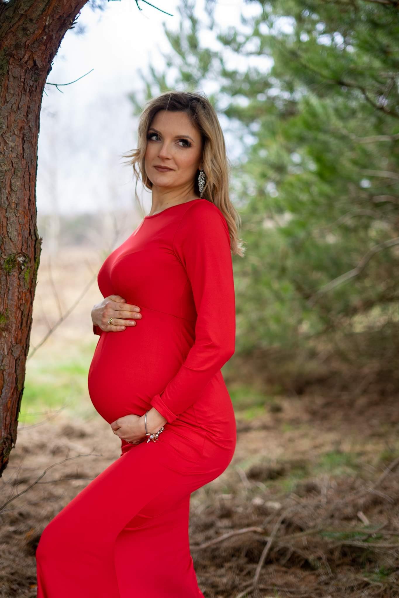Kobieta w czerwonej sukni ciążowej, opierająca się o drzewo na tle leśnym, z rękami na brzuchu i zamyślonym spojrzeniem.