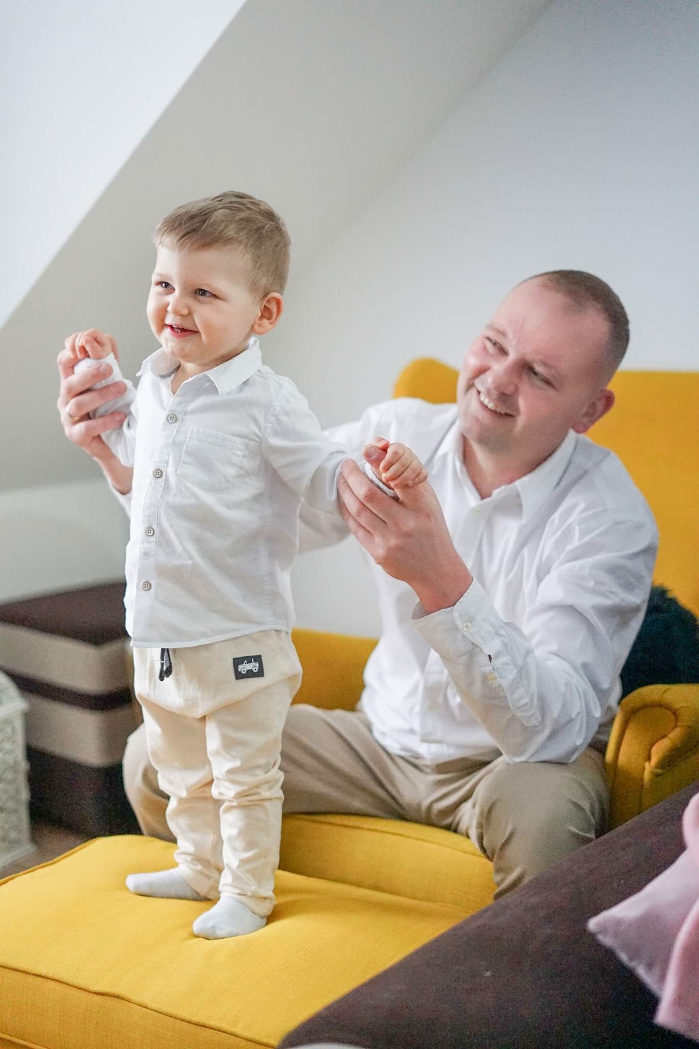 Uśmiechnięty ojciec pomaga synkowi w białej koszuli i beżowych spodniach wstać z żółtej pufy. Chłopiec patrzy w lewo z radością.