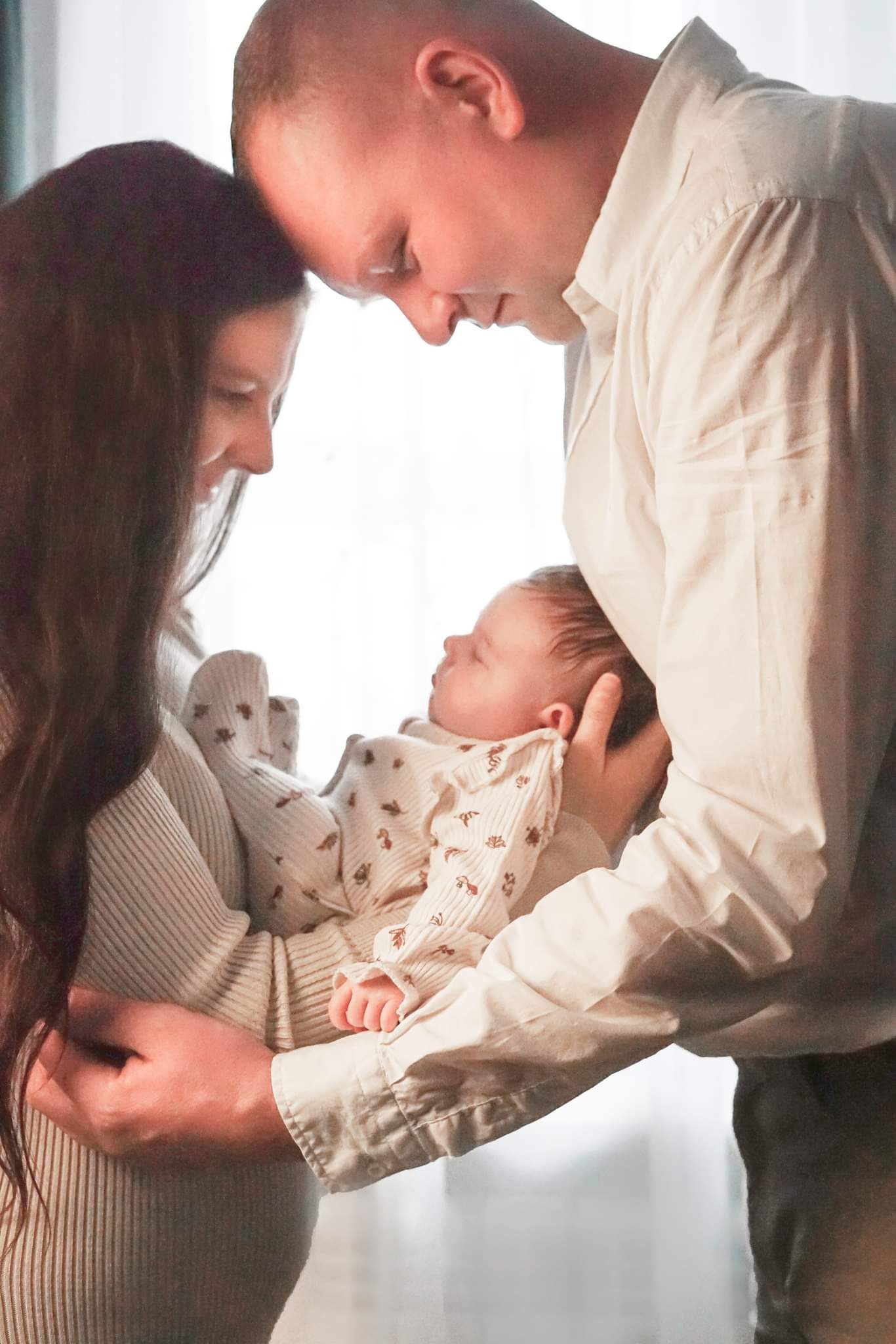 Rodzice trzymający śpiącego noworodka w ramionach, matka patrzy z uśmiechem, głowy rodziców dotykają się.