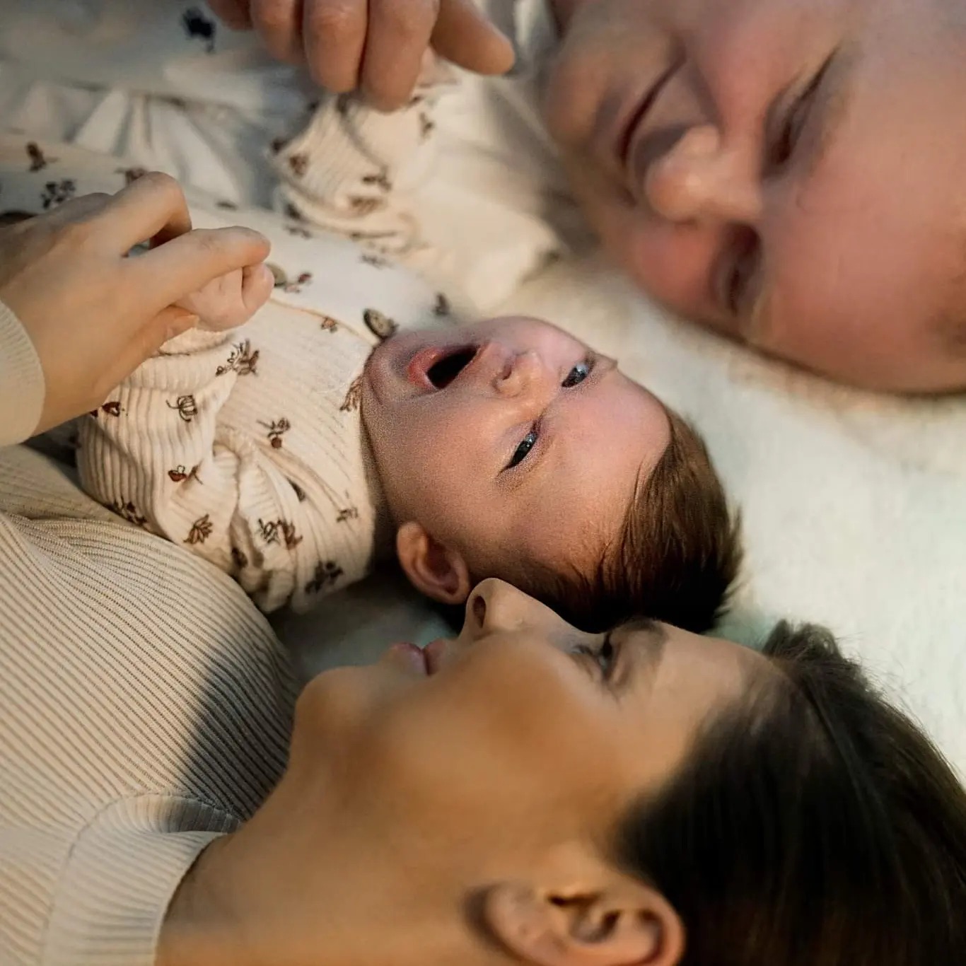 Rodzice leżący obok noworodka z otwartymi ustami, ujęcie z góry, delikatne oświetlenie.