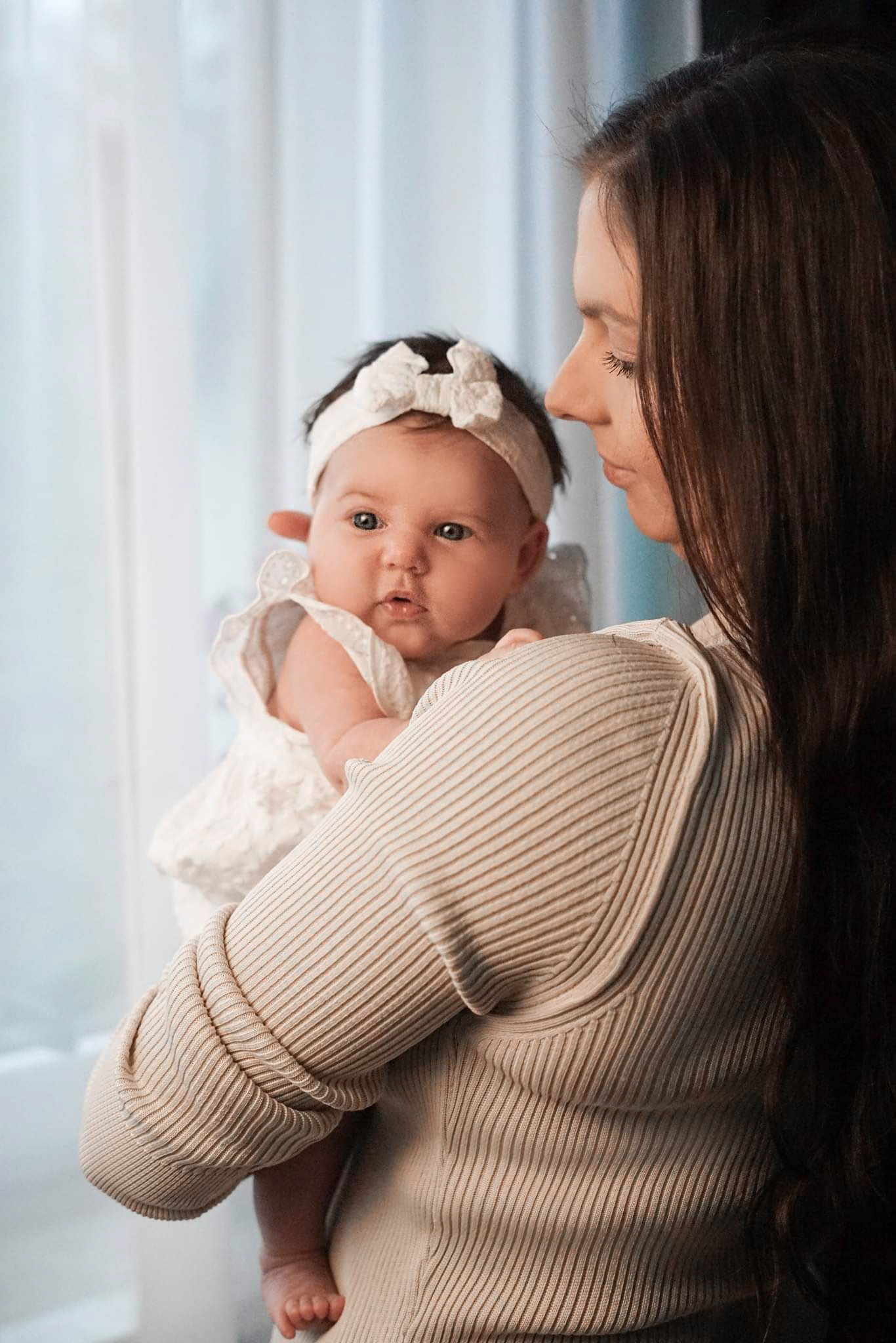 Matka trzyma noworodka w białej sukience i opasce z kokardką na tle okna z delikatną firanką.
