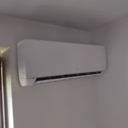 Biały klimatyzator AUX zamontowany na białej ścianie w rogu pokoju, widoczne żaluzje w dolnej części urządzenia.