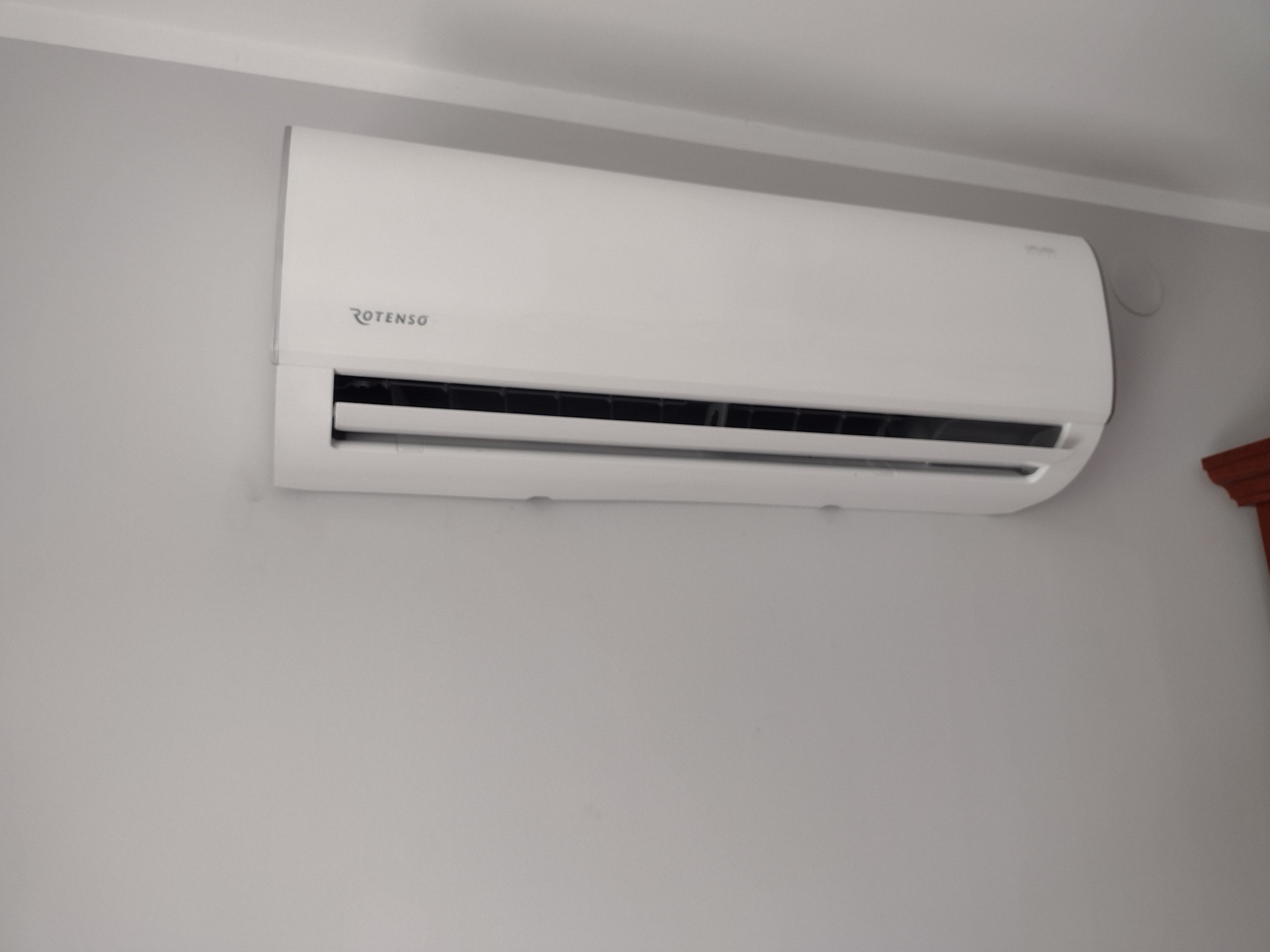 Zamontowany na szarej ścianie klimatyzator Rotenso w jasnym odcieniu bieli, z widocznym fragmentem drewnianej dekoracji w prawym górnym rogu.