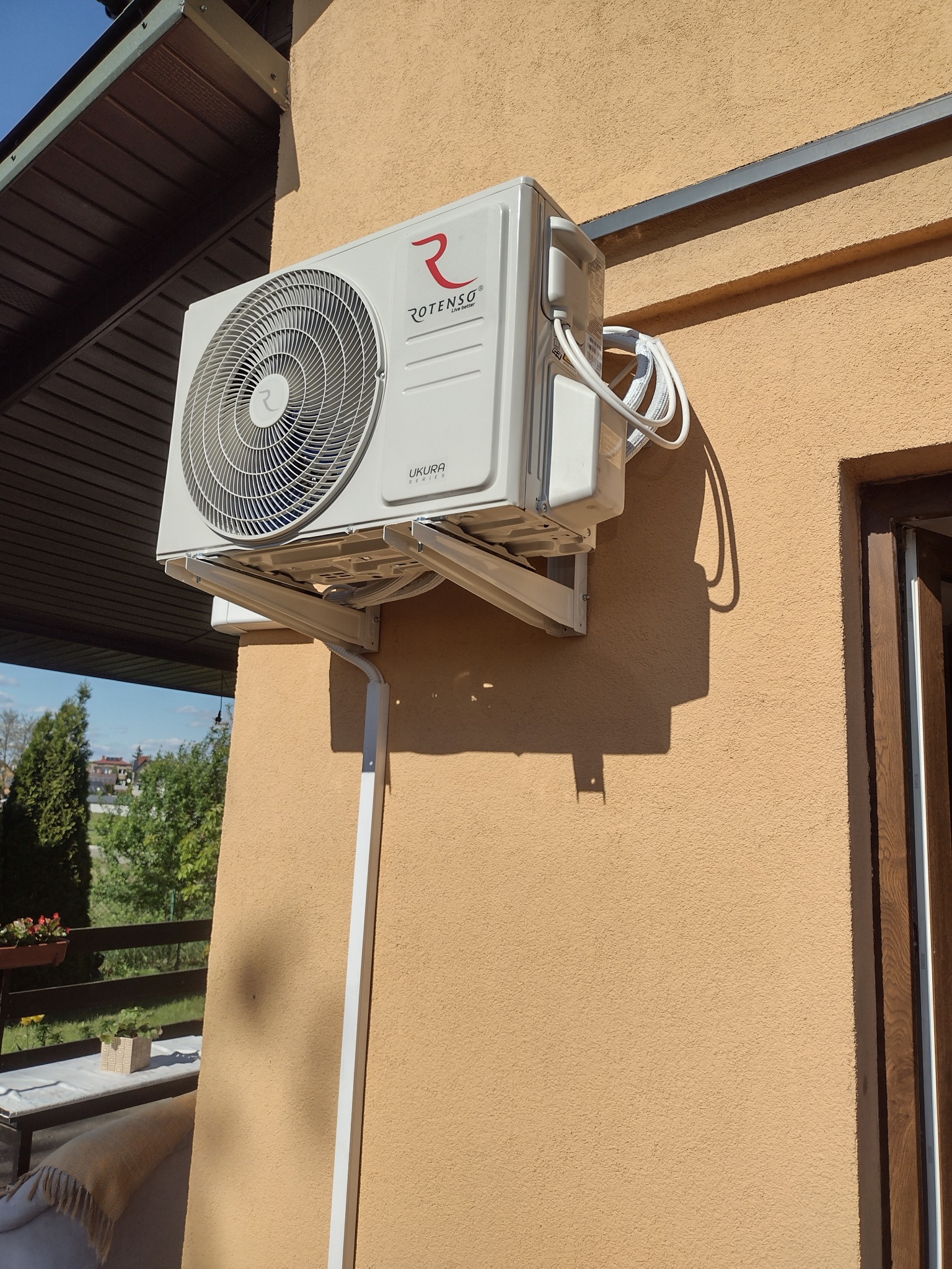 Biała jednostka zewnętrzna klimatyzatora Rotenso Ukura zamontowana na elewacji domu o beżowej barwie, widoczne przewody i rurka odprowadzająca skropliny, w tle zieleń drzew i fragment balkonu.
