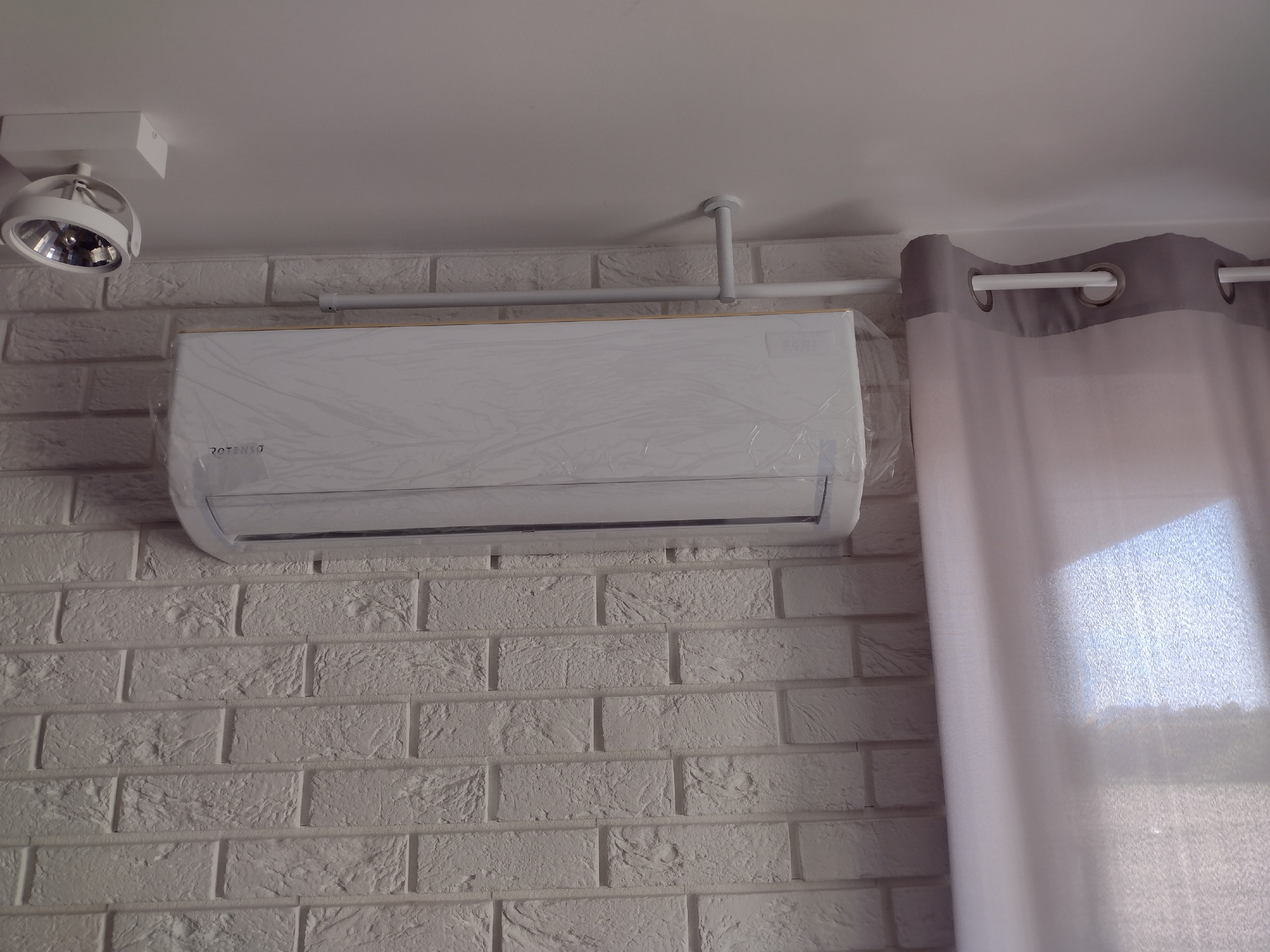 Nowy klimatyzator, jeszcze w folii ochronnej, zamontowany na ścianie z białej cegły w pomieszczeniu z szarymi zasłonami i punktowym oświetleniem.