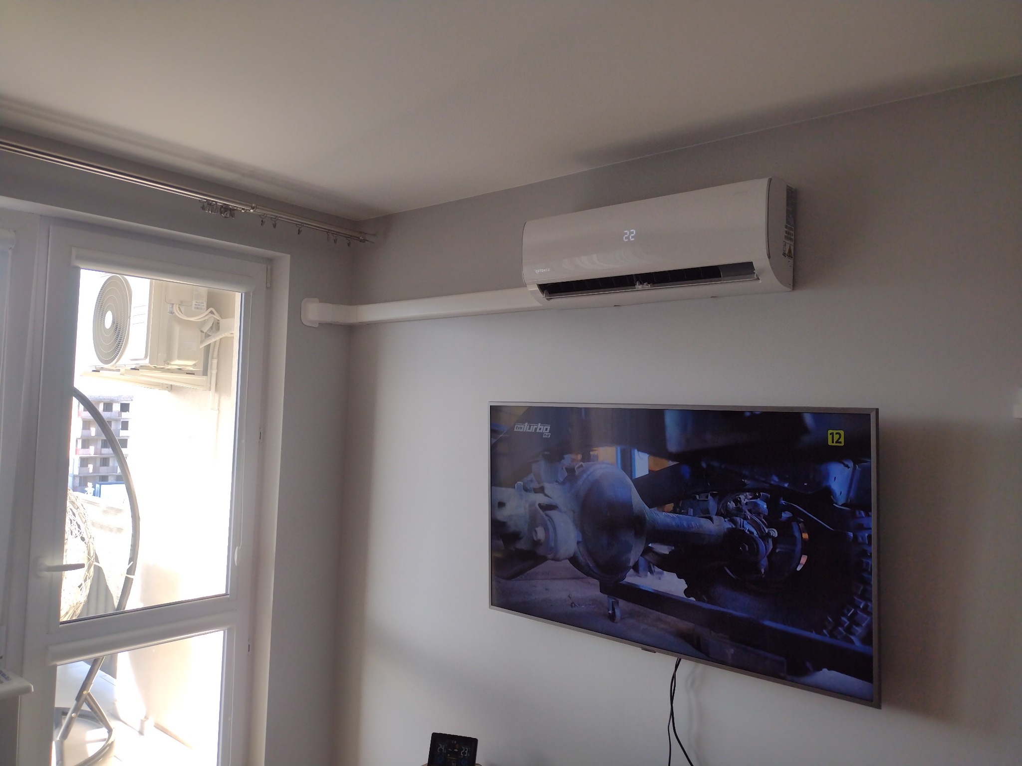 Biały klimatyzator ścienny z widocznym wyświetlaczem temperatury 22 stopnie Celsjusza, zamontowany w pokoju z telewizorem i balkonem.