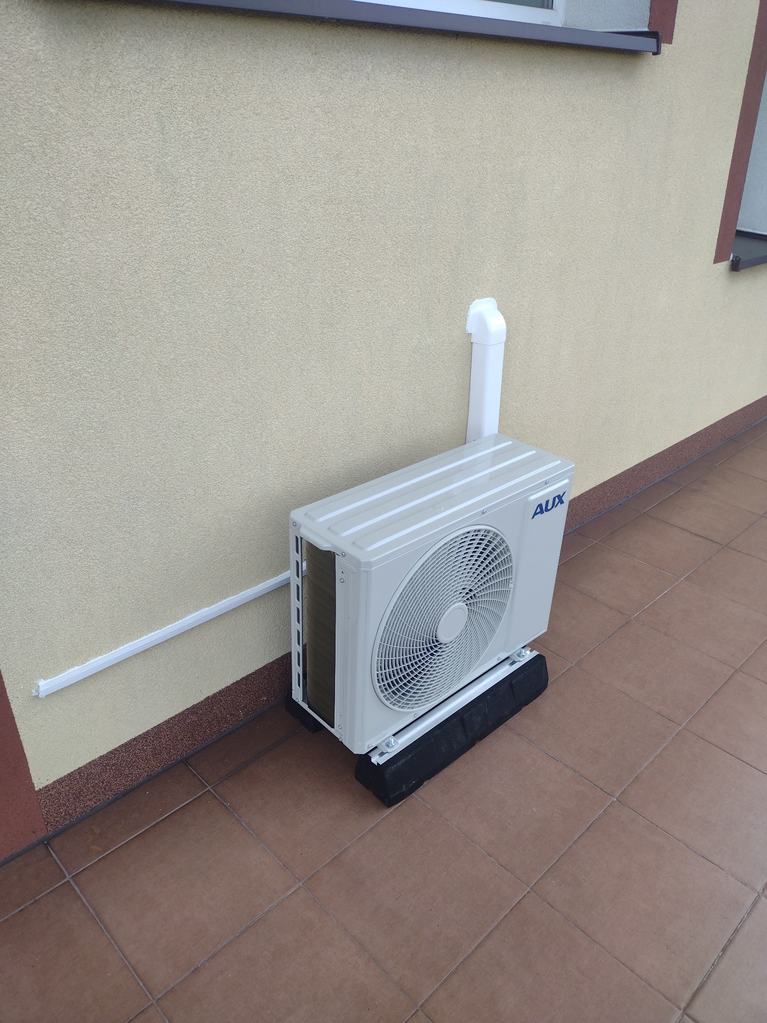 Zewnętrzna jednostka klimatyzacji AUX zamontowana na balkonie, widoczne orurowanie przy ścianie budynku, brązowe płytki podłogowe.
