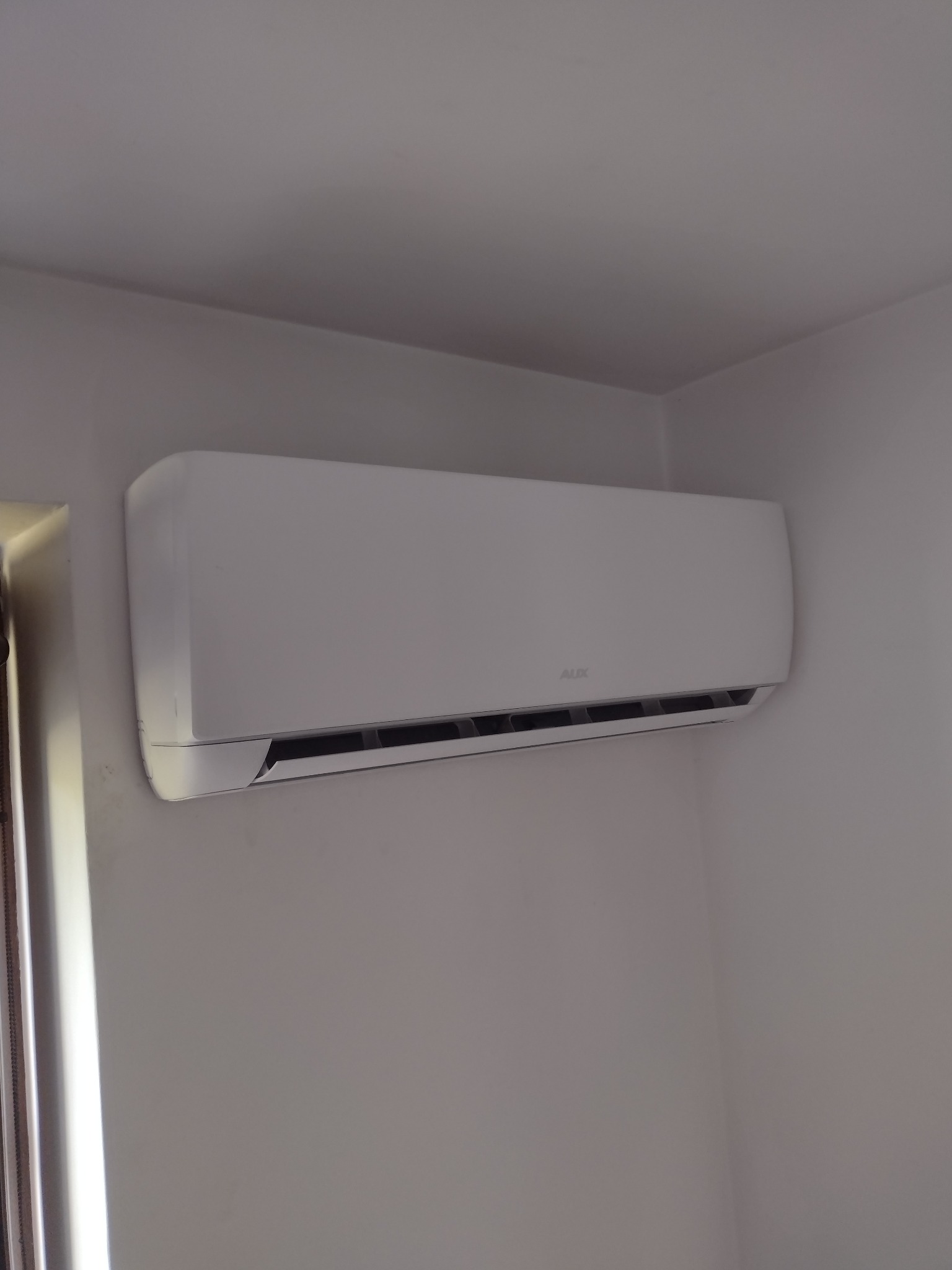 Biały klimatyzator AUX zamontowany na białej ścianie w rogu pokoju, widoczne żaluzje w dolnej części urządzenia.