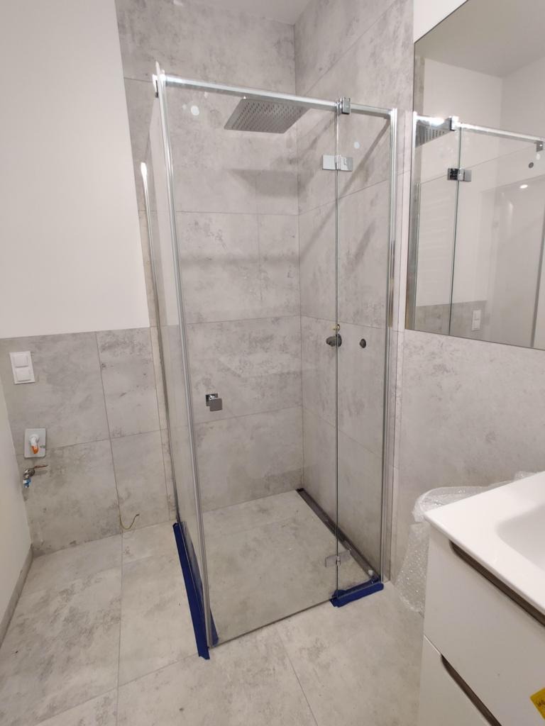 Nowoczesna kabina prysznicowa typu walk-in z deszczownicą, wykończona szarymi płytkami imitującymi beton. Widoczne zabezpieczenia na dole kabiny.