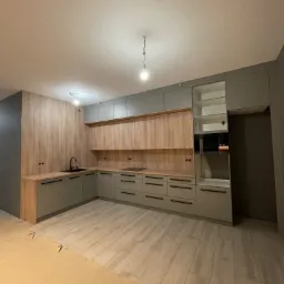 Jasnoszara kuchnia w nowym mieszkaniu z drewnianymi akcentami, czarnym zlewem i płytą grzewczą, częściowo zakryta folią ochronną na podłodze.