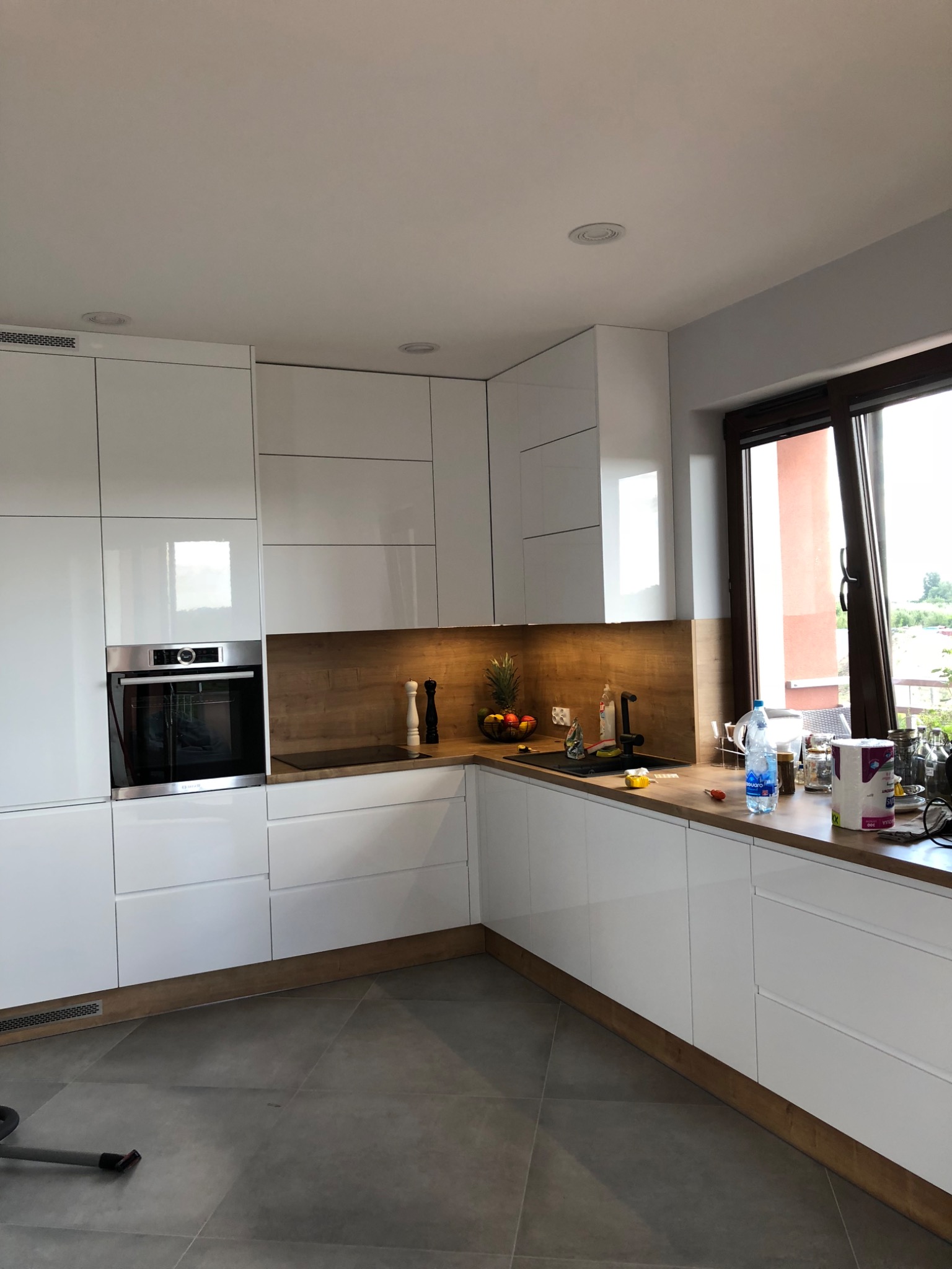 Nowoczesna, biała kuchnia z drewnianym blatem i czarnym zlewem, oświetlona światłem dziennym wpadającym przez okno, z widocznymi akcesoriami kuchennymi i elementami dekoracyjnymi na blacie.