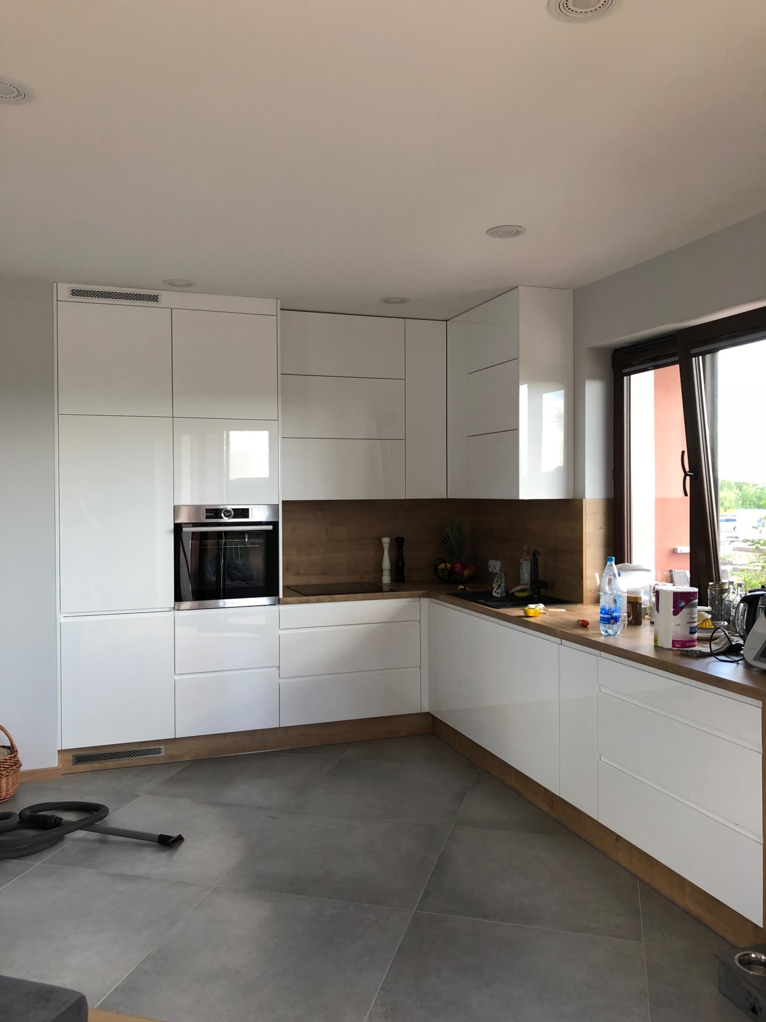 Nowoczesna kuchnia z białymi, lakierowanymi frontami szafek, drewnianym blatem i brązowym panelem nad blatem, widok na okno z widokiem na zieleń, na blacie owoce i akcesoria kuchenne, na podłodze...