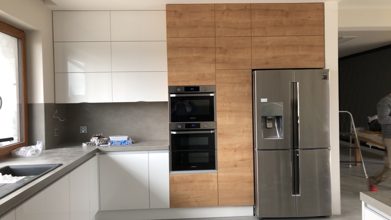 Nowoczesna kuchnia w trakcie montażu: białe szafki kontrastują z drewnopodobnymi, zabudowane piekarniki i srebrna lodówka side-by-side, widoczne elementy instalacji elektrycznej.