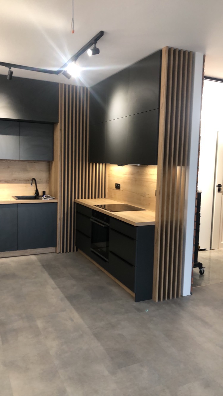 Nowoczesna kuchnia z ciemnymi szafkami i blatem w odcieniu drewna, oddzielona od reszty pomieszczenia drewnianymi lamelami, oświetlenie punktowe na szynie.