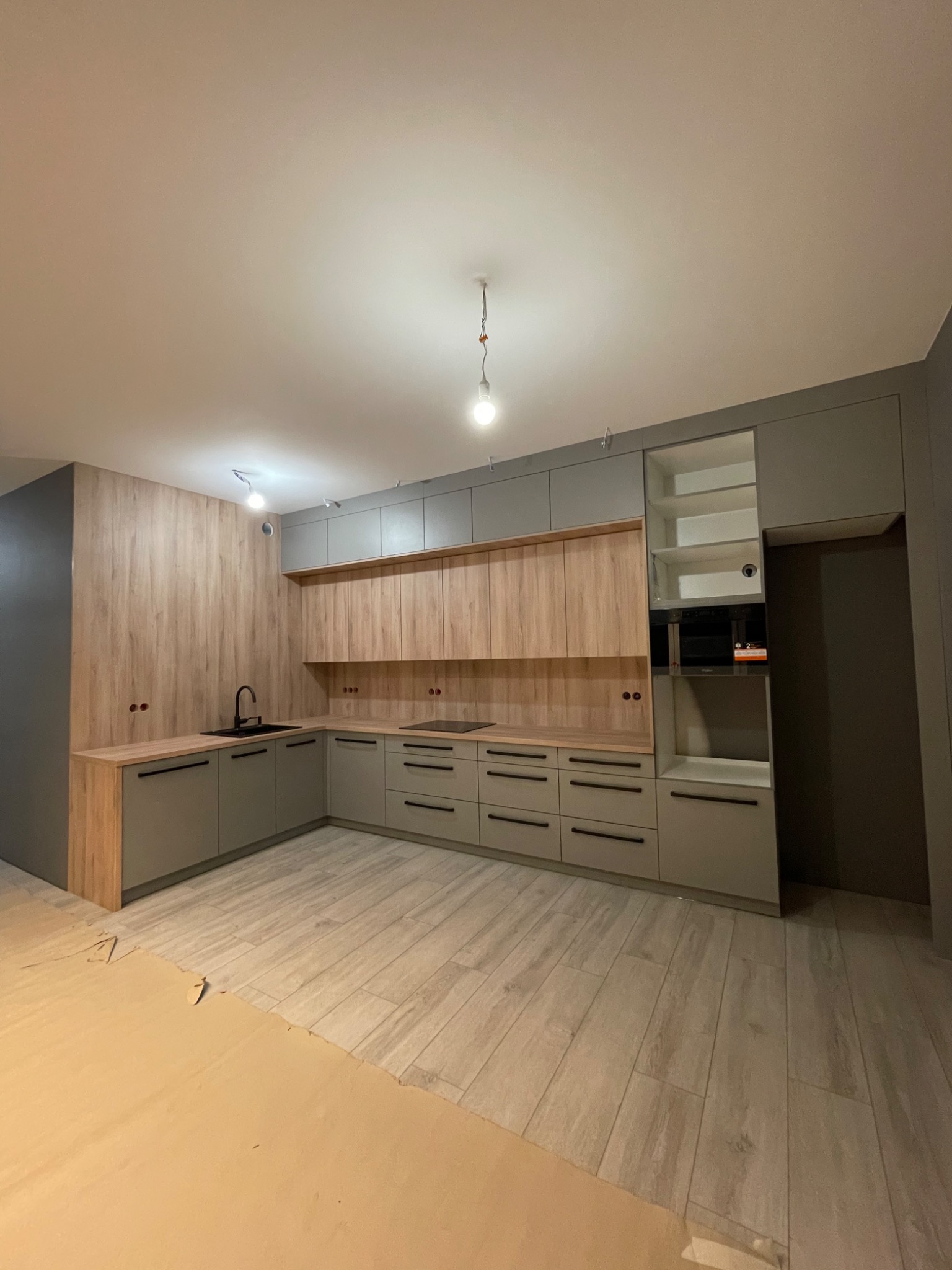 Jasnoszara kuchnia w nowym mieszkaniu z drewnianymi akcentami, czarnym zlewem i płytą grzewczą, częściowo zakryta folią ochronną na podłodze.