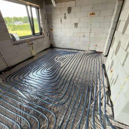 NIK INSTAL MARCIN BAJEWSKI - Instalacja ogrzewania podłogowego z rur w kształcie pętli na folii izolacyjnej w surowym wnętrzu z oknem i widocznymi przewodami elektrycznymi na ścianach z betonu komórkowego.