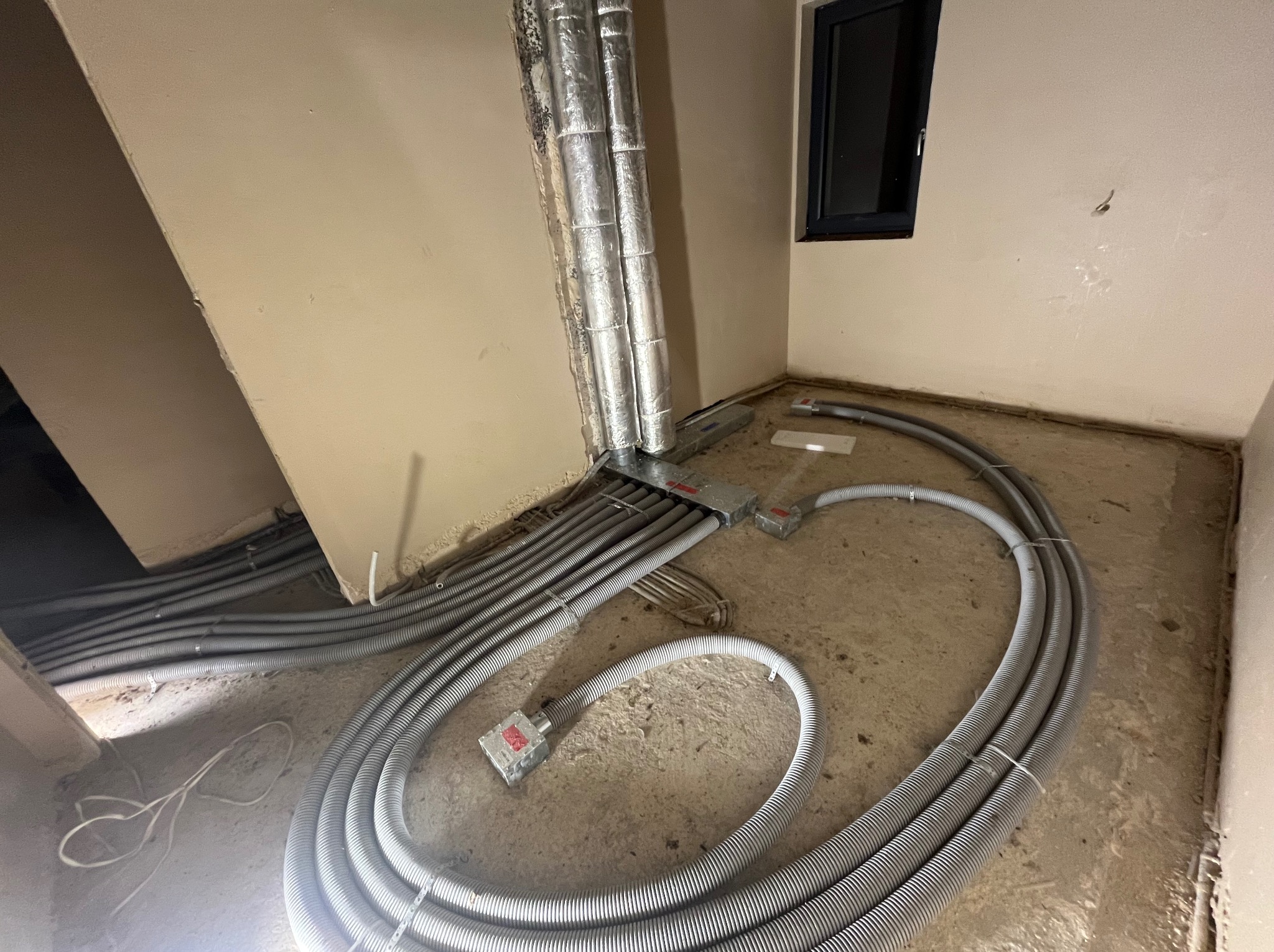 Szare, karbowane rury instalacyjne ułożone na surowym podłożu w rogu pomieszczenia z oknem. Rury prowadzą do pionowych, zaizolowanych przewodów.