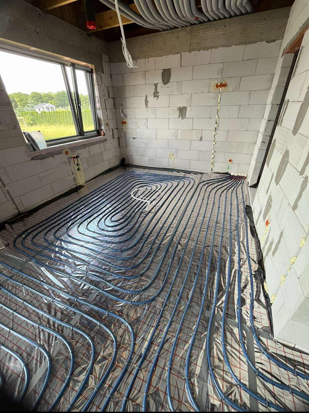 Instalacja ogrzewania podłogowego z rur w kształcie pętli na folii izolacyjnej w surowym wnętrzu z oknem i widocznymi przewodami elektrycznymi na ścianach z betonu komórkowego.