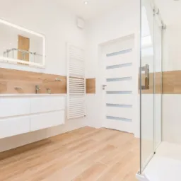 Nowoczesna łazienka z białymi płytkami, drewnopodobnymi akcentami, podświetlanym lustrem, przeszkloną kabiną prysznicową i podłogą z paneli imitujących drewno.