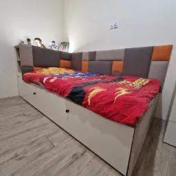 Łóżko dziecięce z szufladami i tapicerowanym wezgłowiem w odcieniach szarości i pomarańczy, widoczne z boku w pomieszczeniu w Babiaku.
