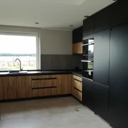 Nowoczesna kuchnia z drewnianymi frontami szafek dolnych i czarnymi szafkami górnymi, widok na okno z widokiem na pole, jasna podłoga.