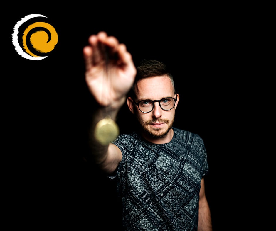 Hipnoterapeuta w okularach, z brodą, ubrany w koszulkę we wzory, trzymający wahadełko na czarnym tle, z logo firmy w lewym górnym rogu.