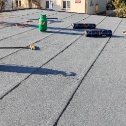 Płaski dach pokryty papą termozgrzewalną, widoczne zgrzewy, butle z gazem propan-butan, szczotka dekarska i częściowo widoczna drabina oparta o krawędź dachu.