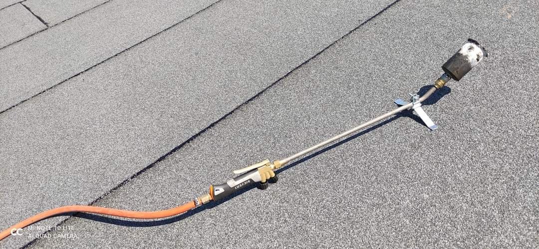 Palnik dekarski z pomarańczowym wężem leżący na pokryciu z papy bitumicznej z widocznymi łączeniami.