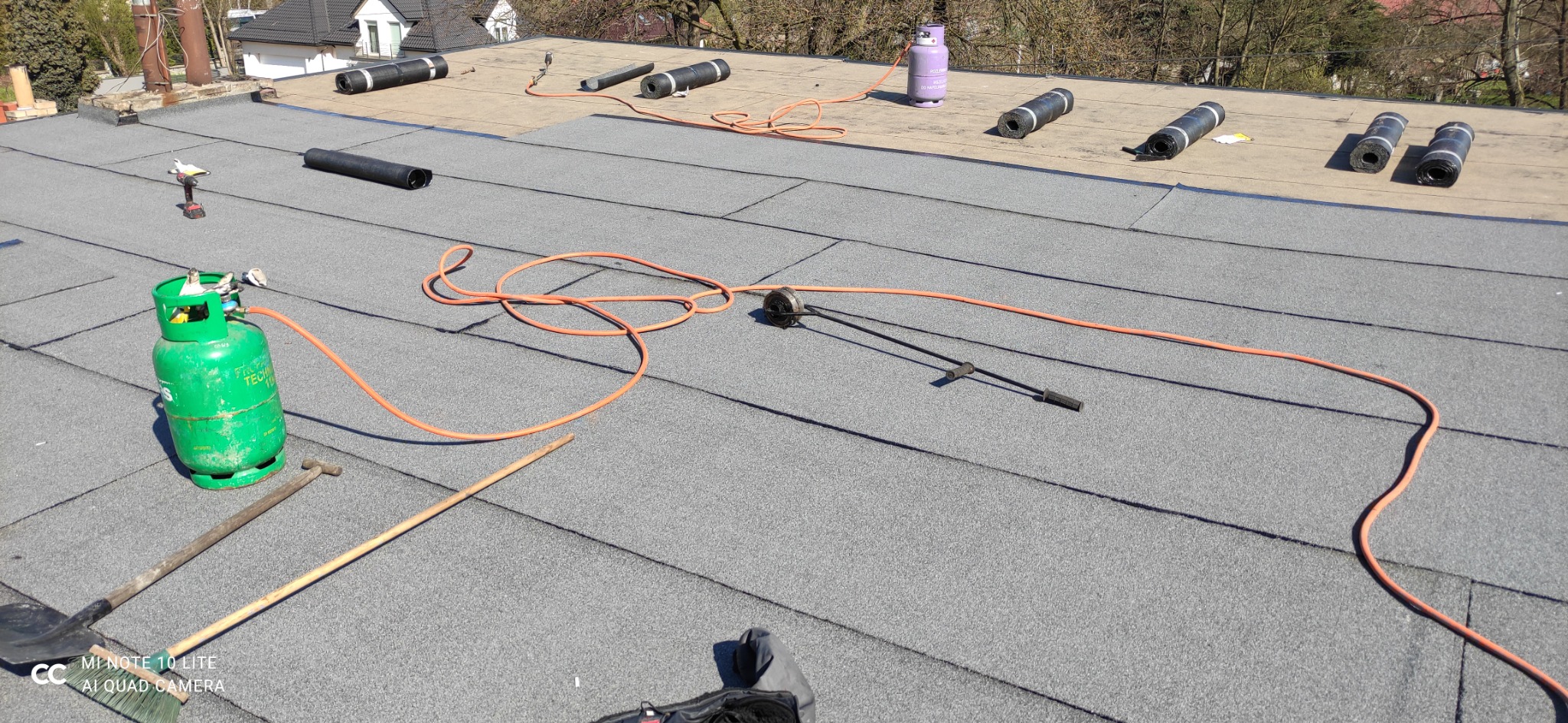 Fragment dachu płaskiego z papy termozgrzewalnej w trakcie prac, widoczne butle gazowe, rolki papy i narzędzia dekarskie, w tle domy.