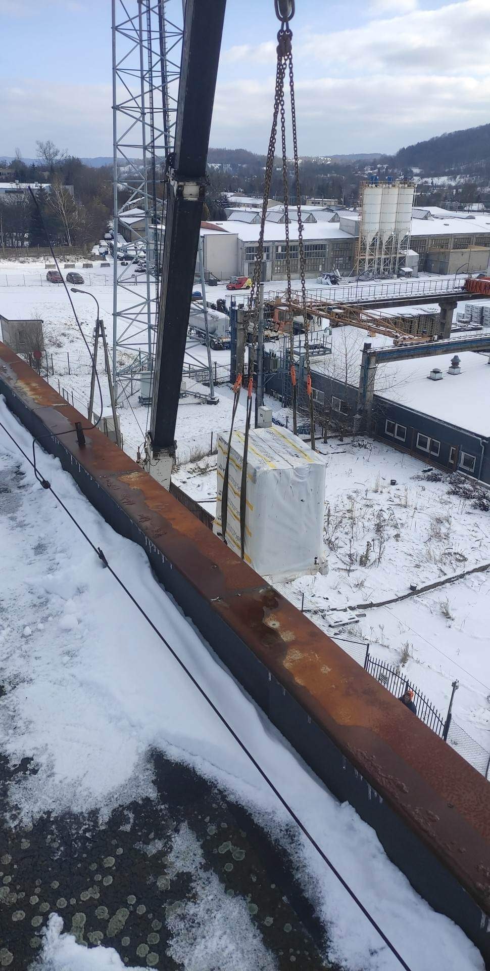 Żuraw podnosi pakiet materiałów budowlanych na tle zimowego krajobrazu z budynkami pokrytymi śniegiem.