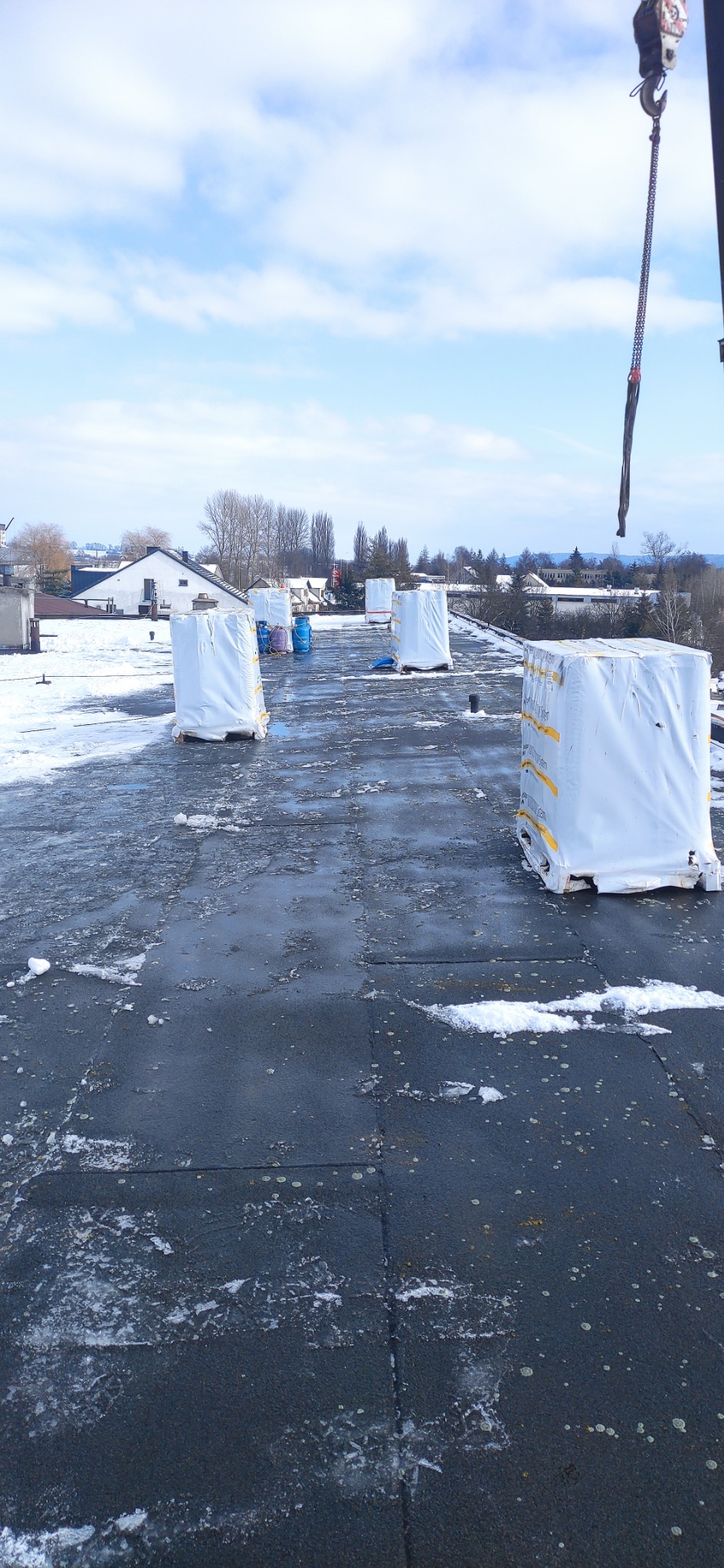 Płaski dach pokryty bitumiczną papą, częściowo ośnieżony, z przygotowanymi do montażu, owiniętymi folią pakietami materiałów budowlanych, widoczny dźwig i zabudowania w tle.