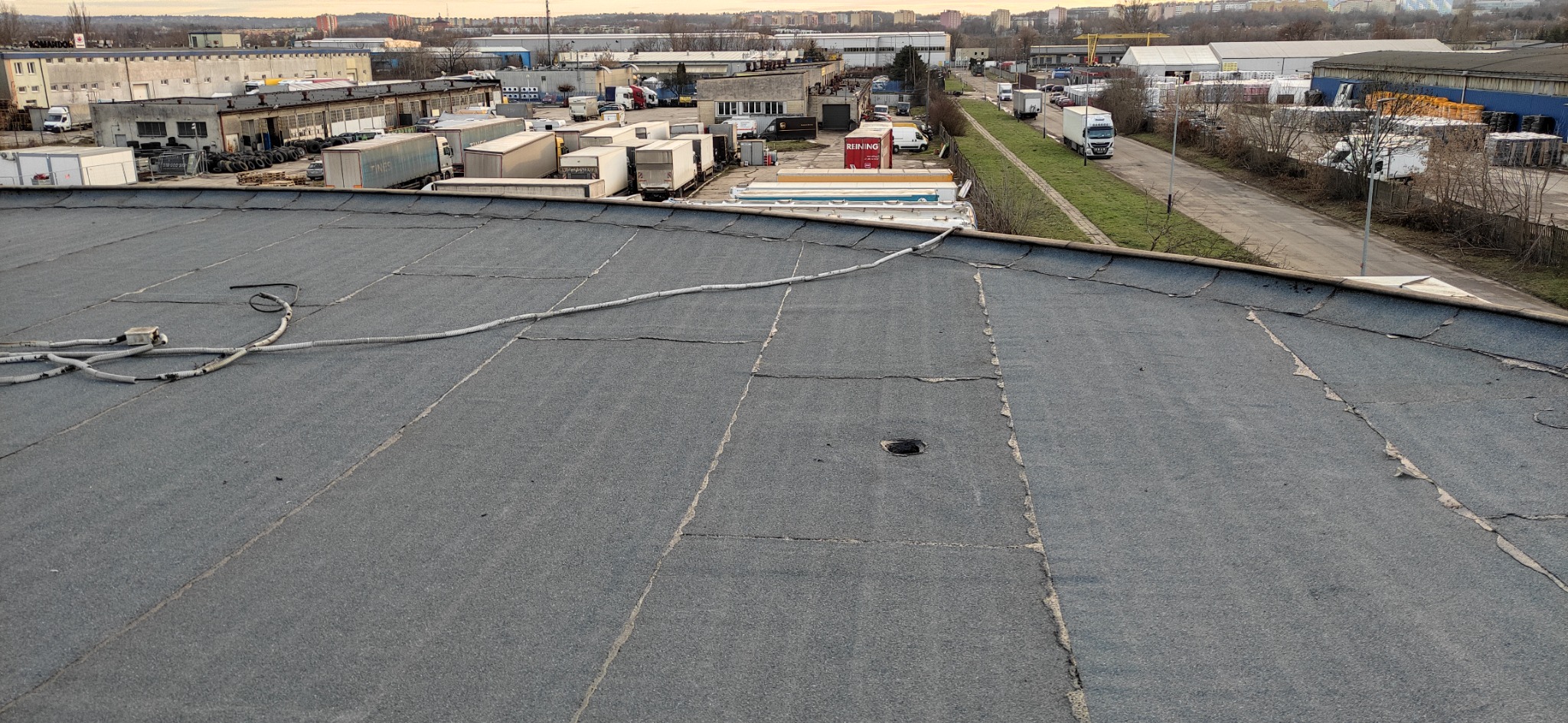 Płaski, szary dach z papy na budynku przemysłowym, widoczne pęknięcia i uszkodzenia, z przewodami elektrycznymi i otworem w poszyciu, w tle zabudowania przemysłowe i krajobraz miejski.
