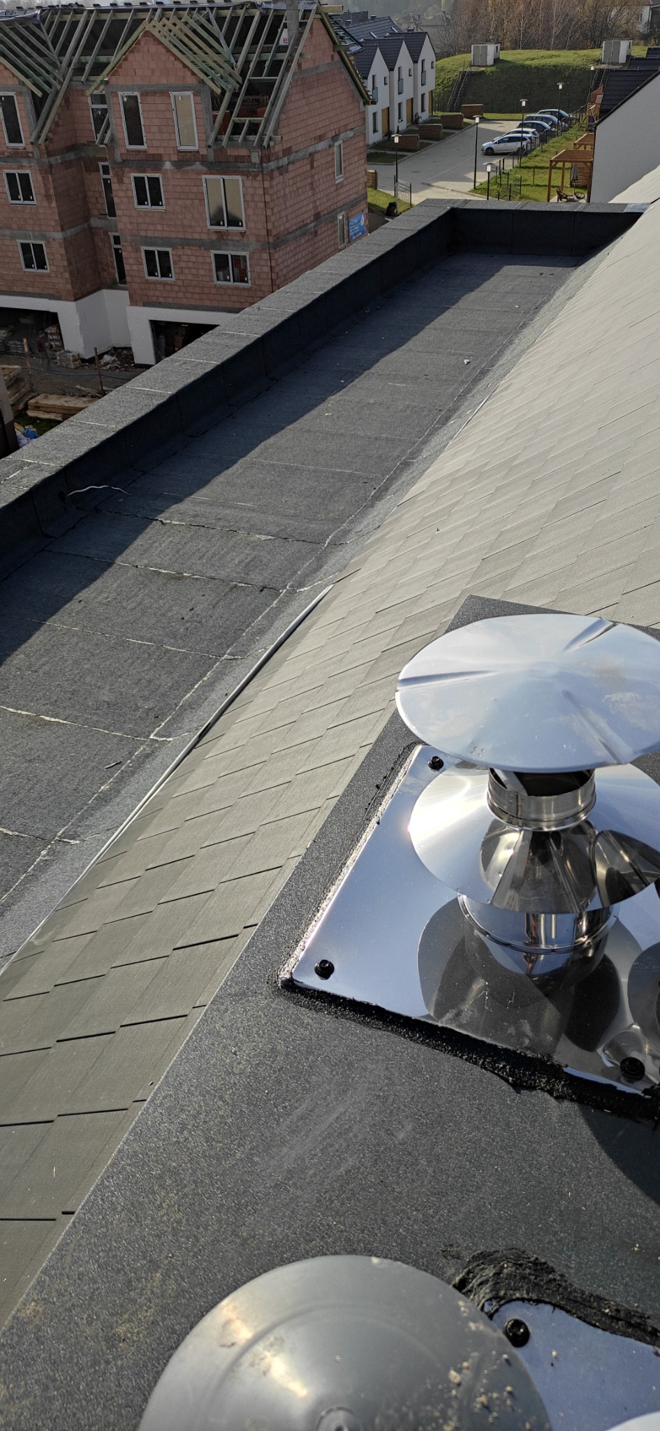 Szczegółowe ujęcie dachu z papy z widocznym kominkiem wentylacyjnym z blachy nierdzewnej, w tle budynek w stanie surowym.