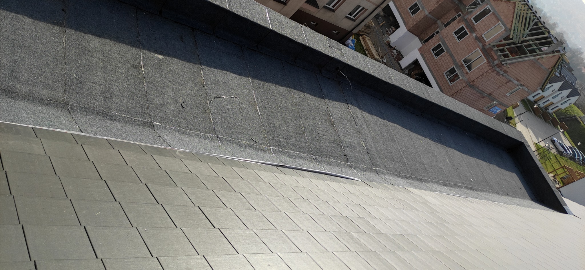 Fragment dachu z papy termozgrzewalnej z widocznymi łączeniami i fragment dachu pokrytego szarą dachówką bitumiczną na tle budynków mieszkalnych i placu budowy.