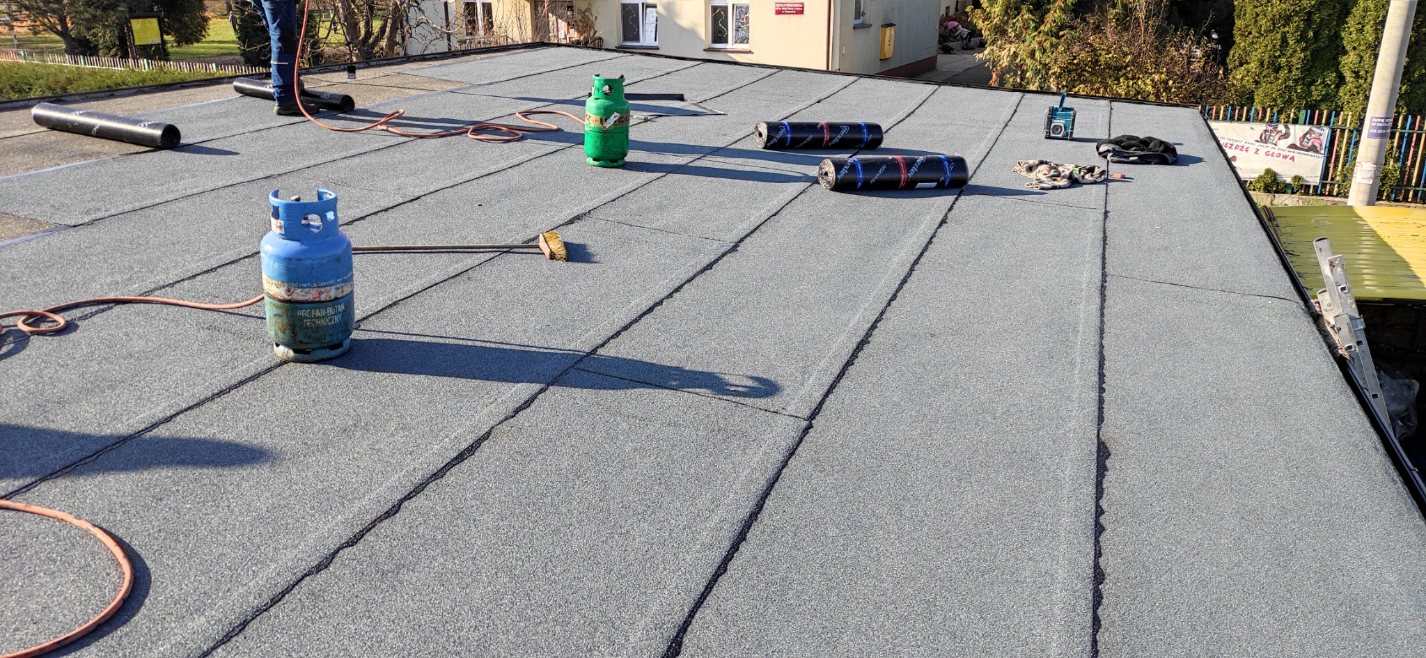 Płaski dach pokryty papą termozgrzewalną, widoczne zgrzewy, butle z gazem propan-butan, szczotka dekarska i częściowo widoczna drabina oparta o krawędź dachu.