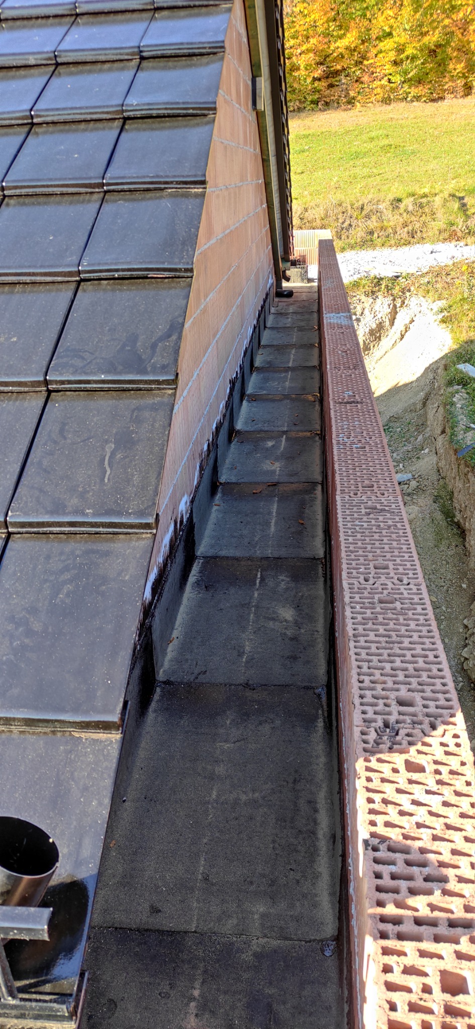 Fragment dachu z ciemnej dachówki ceramicznej, widoczny mur z cegły kratówki oraz rynna z czarną papą wewnątrz, na tle zieleni i drzew.